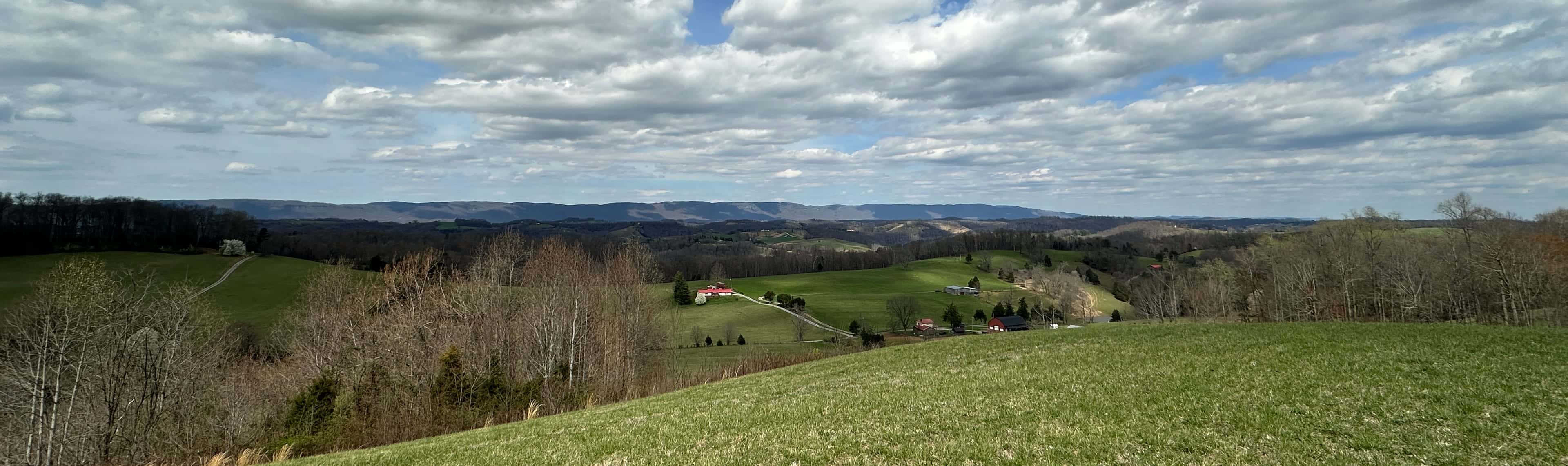 Rolling farmland under a wide sky of broken cloud, a red-roofed farmhouse and outbuildings in the middle distance and long ridges along the horizon