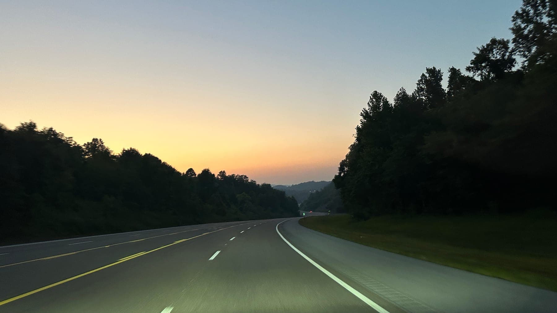 A four-lane highway curving through wooded hills at dusk, the last gold of the sunset along the horizon