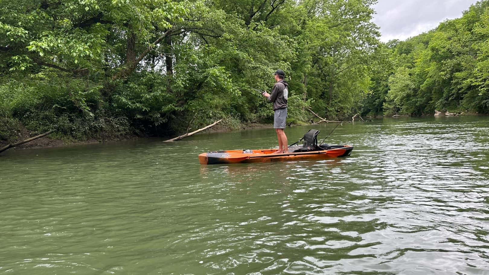 A person standing upright on an orange fishing kayak in the middle of a calm river, casting toward the far bank