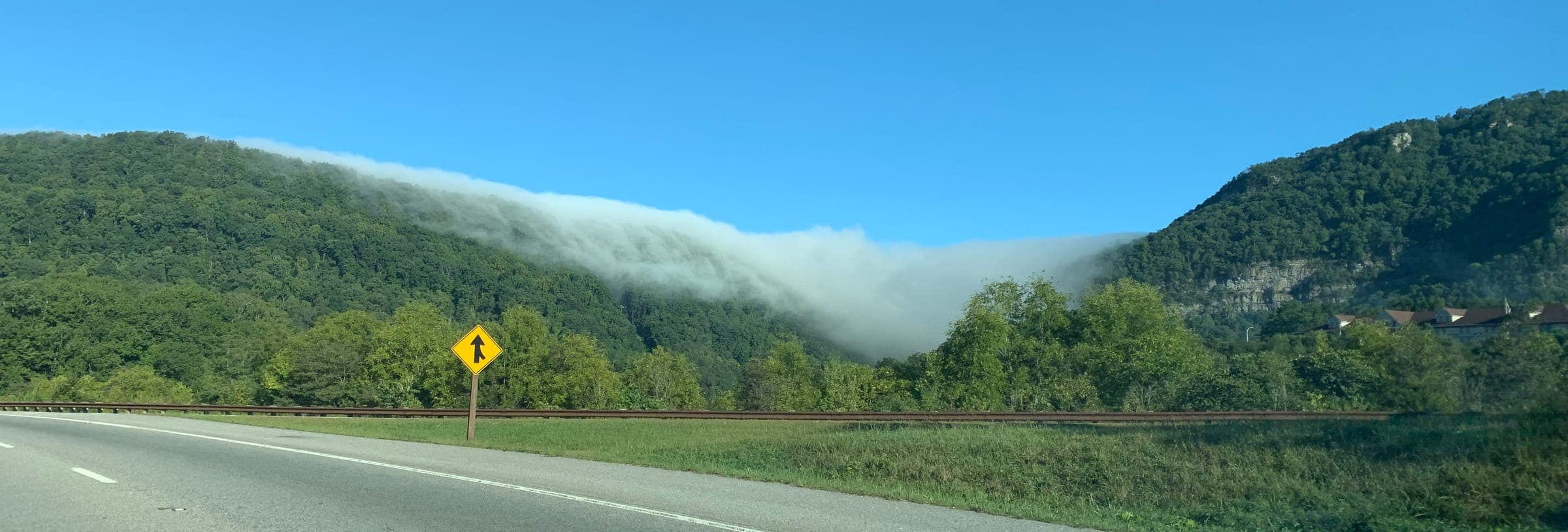 A low cloud spilling over the crest of a forested mountain ridge under a clear blue sky, with a two-lane road and a rail line running along the foot of the slope