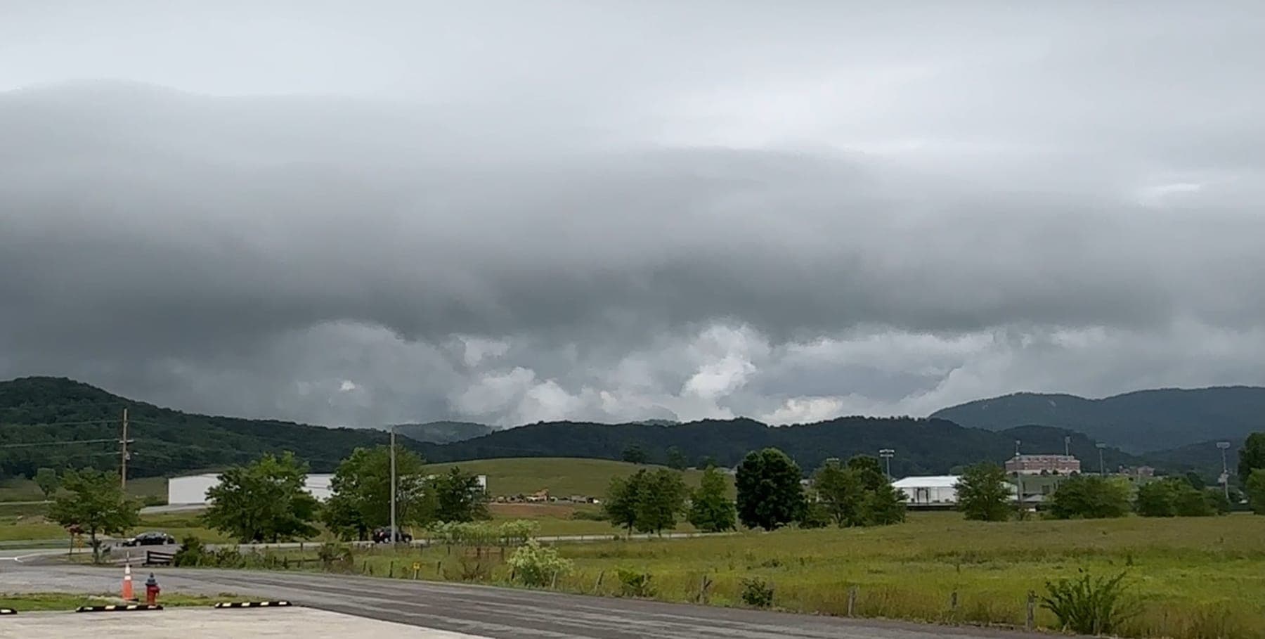 A wide valley under a heavy shelf of storm cloud, with wooded ridges along the far side and a large brick college building among the fields below