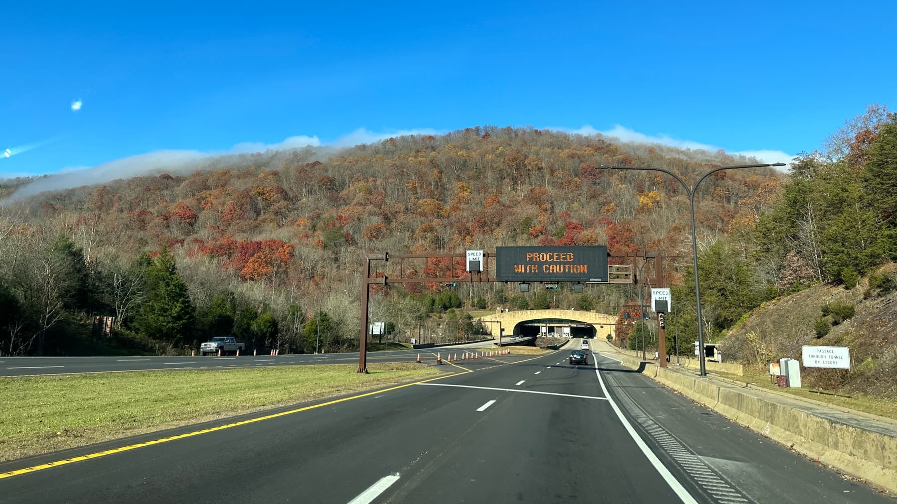 A divided highway running toward a tunnel portal cut into a wooded mountainside in fall color, with an overhead sign reading PROCEED WITH CAUTION