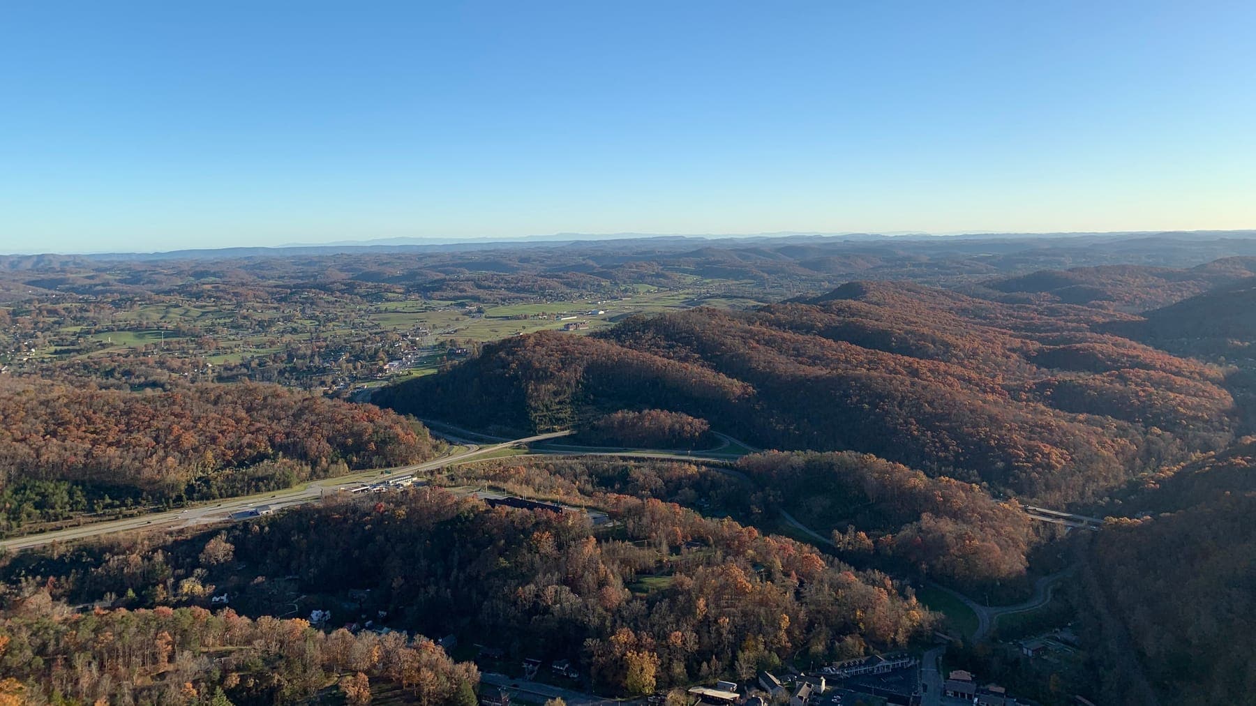 A valley seen from high on a ridge in late fall color, with a highway, a scatter of buildings and long blue ridgelines running to the horizon