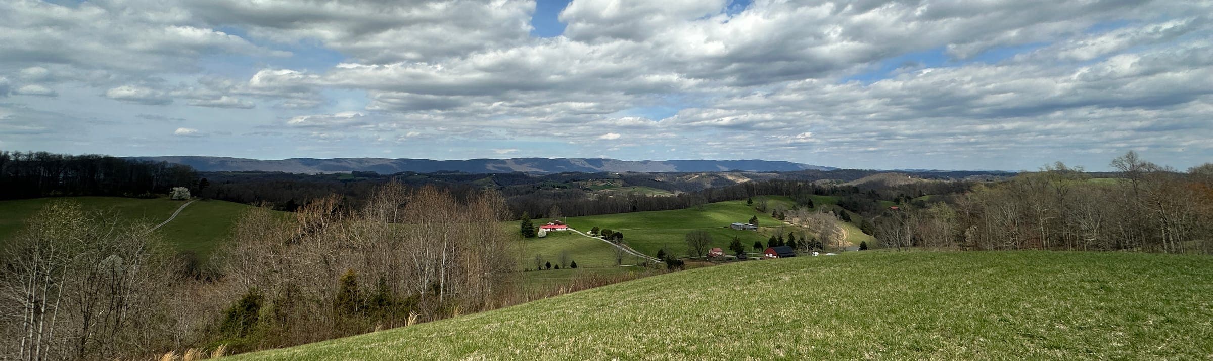 Rolling farmland under a wide sky of broken cloud, a red-roofed farmhouse and outbuildings in the middle distance and long ridges along the horizon
